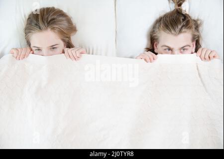 Ein junges Paar im Bett bedeckt die Hälfte ihrer Gesichter mit einer Decke, die sich hinter einer Decke versteckt. Das Konzept der familienfreien p Stockfoto