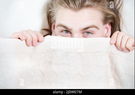 Ein junger attraktiver Mann guckt unter den Decken, die sich hinter ihnen verstecken, mit Angst heraus, wenn er aufwacht. Das Konzept der männlichen Angst Stockfoto