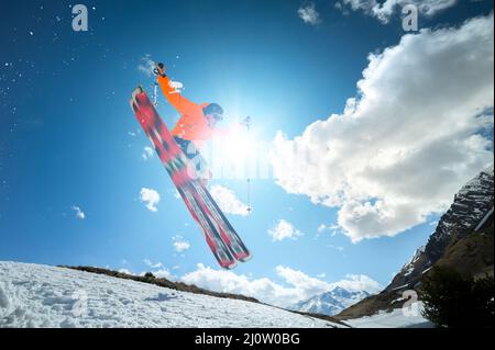 Ein junger stilvoller Mann mit Sonnenbrille und Mütze macht einen Trick, wenn er mit einem Kicker Schnee gegen den blauen Himmel und die Sonne springt o Stockfoto