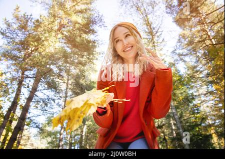 Attraktives weißes kaukasisches Mädchen in rotem Polto und orangefarbenem Hut wird mit einem Blumenstrauß von Blättern in ihren Händen gegen den Backgr verwechselt Stockfoto