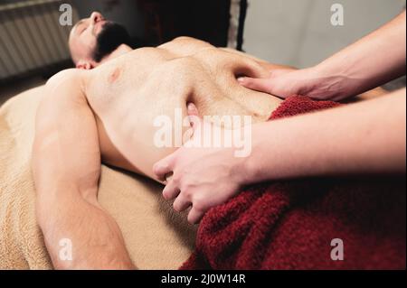 Der männliche Masseur massiert das Thoraxdiaphragma eines muskulösen männlichen Athleten in einem Massageraum mit dunkler Beleuchtung Stockfoto