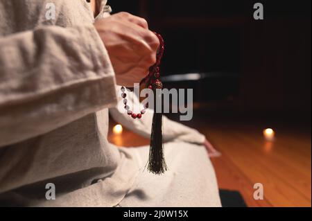 Nahaufnahme Mann in Kleidung für Praxis und Meditation sitzt in einer Lotushaltung und hält roten Rosenkranz, um die Aufmerksamkeit in einem Wald zu konzentrieren Stockfoto