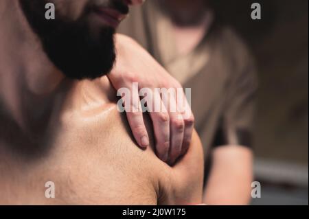 Der männliche Masseur aus der Nähe massiert einen muskulösen männlichen Sportler in einem Raum mit kontrastreicher Beleuchtung mit einer sportlichen Schultermassage. Bekennen Stockfoto