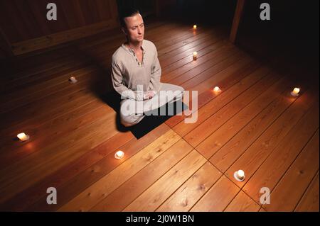 Ein Mann in Kleidung zum Üben und Meditieren sitzt in einer Lotushaltung und hält einen roten Rosenkranz, um die Aufmerksamkeit in einem Holzraum zu konzentrieren Stockfoto