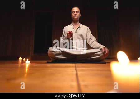 Ein Mann in Kleidung zum Üben und Meditieren sitzt in einer Lotushaltung und hält einen roten Rosenkranz, um die Aufmerksamkeit in einem Holzraum zu konzentrieren Stockfoto