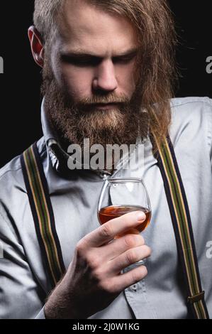 Ein attraktiver Mann mit einem langen Knallbart und einem Schnurrbart, der an der Wand sitzt, schnuppert den Duft eines alkoholischen Getränks in einem Stockfoto