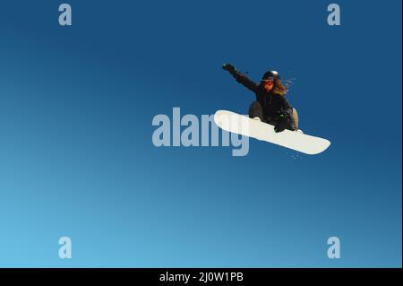 Snowboardermädchen macht einen Trick beim Springen mit einem Griff gegen den blauen Himmel. Blauer Hintergrund isoliert Athlet im Flug Stockfoto