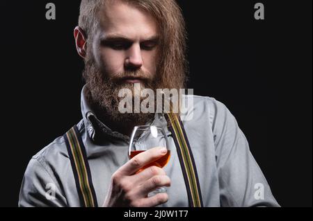 Ein attraktiver Mann mit einem langen Knallbart und einem Schnurrbart, der an der Wand sitzt, schnuppert den Duft eines alkoholischen Getränks in einem Stockfoto