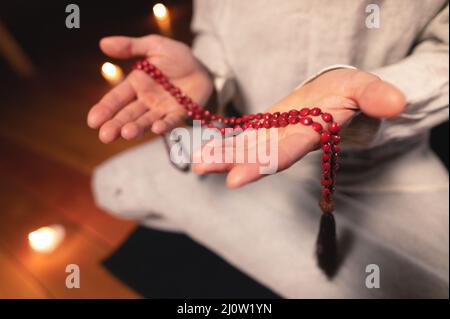 Nahaufnahme Mann in Kleidung für Praxis und Meditation sitzt in einer Lotushaltung und hält roten Rosenkranz, um die Aufmerksamkeit in einem Wald zu konzentrieren Stockfoto