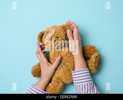 Weibliche Hand hält einen braunen Teddybär und klebt einen medizinischen Klebstoff-Gips auf blauem Hintergrund, Tram-Behandlung Stockfoto