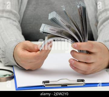 Ein Stapel von hundert Dollar Scheinen in der Hand einer Frau und eine hölzerne Lupe auf einem weißen Tisch. Budgetanalyse, Einkommen und Stockfoto