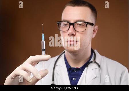 Junger kaukasischer Arzt bereitet sich auf eine Impfung mit einer Spritze vor. Studio auf braunem Hintergrund. Lassen Sie die Luft aus dem ab Stockfoto