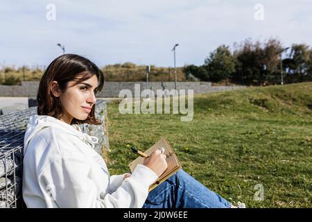 Junge hispanische Studentin, die im öffentlichen Park sitzt und in einem Notizbuch schreibt Stockfoto
