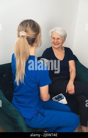 Vertikale Innenaufnahme des Hausbesuchs. Glückliche ältere Frau mit Hyperspannung und ihre junge blonde Betreuerin. Hochwertige Fotos Stockfoto