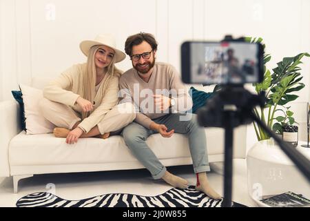 Junge verheiratete lächelndes kaukasisches Paar, das Videoinhalte mit einem Smartphone auf der Couch im Innenbereich macht. Hochwertige Fotos Stockfoto