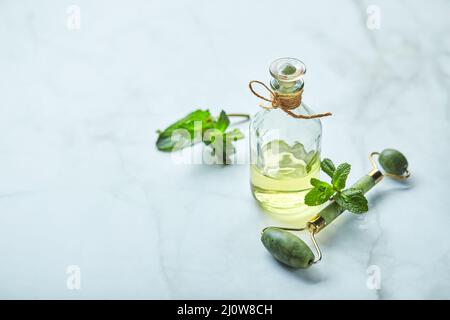 Flasche ätherisches Öl aus Minze und grünen Blättern und Jade-Massagerolle für das Gesicht. Natürliche Bio-Inhaltsstoffe für Kosmetika Stockfoto