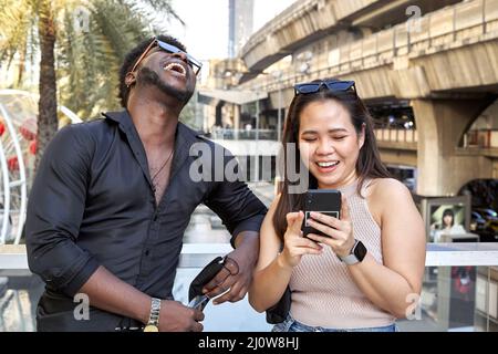 Multirassische Freunde lachen und nutzen das Handy neben einem Einkaufszentrum Stockfoto