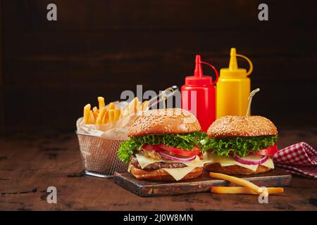 Zwei hausgemachte Burger mit Rind-, Käse- und Zwiebelmarmelade auf einem Holzbrett, Pommes in einem Metallkorb und Saucen. Fast Food Konz Stockfoto