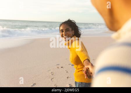 Glückliche junge Birazialfrau, die ihren Freund zieht, während sie die Hand am Strand während des Sonnenuntergangs hält Stockfoto