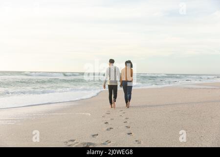 Lange Rückansicht des Biracial Paares beim Gehen, während man Fußabdrücke auf Sand am Strand hinterlässt Stockfoto