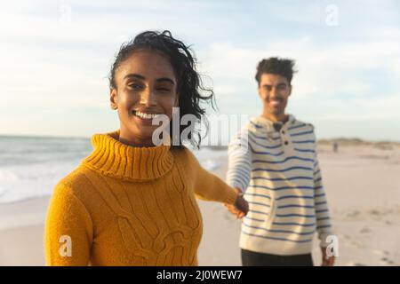 Porträt einer lächelnden jungen Birazialfrau, die Hand hält und einen Freund hinter ihr am Strand steht Stockfoto