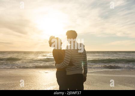 Romantisches Biracial Paar umarmt, während am Strand gegen Himmel während des Sonnenuntergangs stehen Stockfoto