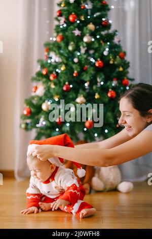 Mutter setzt Weihnachtsmütze auf ihre kleine Tochter Vor einem Weihnachtsbaum Stockfoto