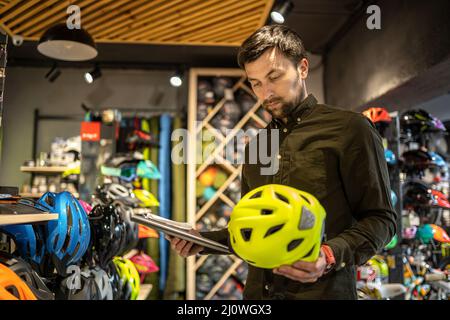 Bike Shop Manager prüft Helmpreisinformationen auf Tablet, Verkäufer macht eine Inventur im Sportgeschäft. Thema der Kleinunternehmen Stockfoto