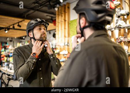 Mann, der Fahrradhelme im Sportgeschäft untersucht und sich im Spiegel ansieht. Neue Sporthelme im Fahrradladen ausprobieren. Mann setzt sich an Stockfoto