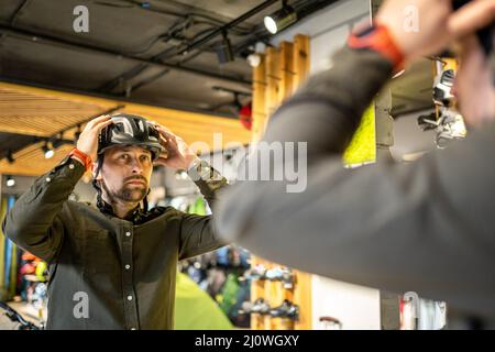 Mann, der Fahrradhelme im Sportgeschäft untersucht und sich im Spiegel ansieht. Neue Sporthelme im Fahrradladen ausprobieren. Mann setzt sich an Stockfoto