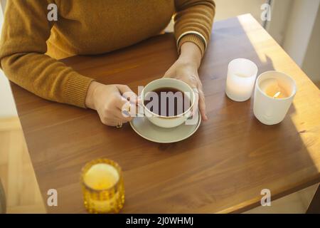 Frau hält eine Tasse heißen Tee, während sie zu Hause mit brennenden Kerzen am Tisch sitzt Stockfoto