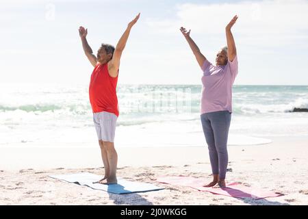 Die ganze Länge des Biracial Senior Paares praktiziert Yoga mit den Armen angehoben am sonnigen Strand gegen den Himmel Stockfoto