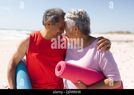 Glückliches Senioren-Paar, das Yogamatten hält, die sich am Strand gegenseitig die Stirn berühren Stockfoto