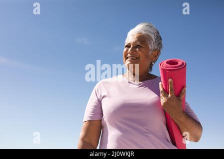 Lächelnde ältere Frau aus der Birazialzeit, die am sonnigen Tag die zusammengerollte Yogamatte am Strand gegen den blauen Himmel hält Stockfoto