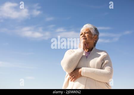 Blick auf die nachdenkliche ältere Birazialfrau mit der Hand am Kinn am Strand vor blauem Himmel Stockfoto