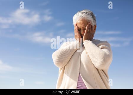 Niedrige Ansicht der besorgten älteren Frau aus der Bira mit den Händen, die das Gesicht am Strand gegen den Himmel bedecken Stockfoto