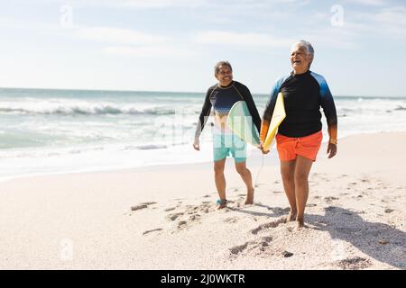 Ein glückliches, multirassisches Seniorenpaar, das mit Surfbrettern auf Sand am sonnigen Strand spazieren geht Stockfoto