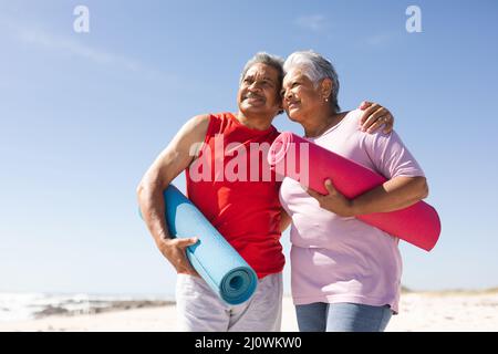 Ein glückliches Paar aus Senioren, das Yogamatten hält und an einem sonnigen Tag den Strand vor dem blauen Himmel sieht Stockfoto