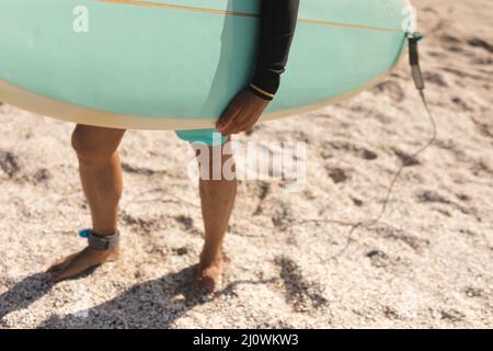 Niedriger Teil eines älteren biracial Mannes, der Surfbrett trägt und barfuß auf Sand am sonnigen Strand steht Stockfoto