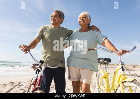 Ein lächelndes, mehrrassiges Ehepaar blickt an sonnigen Tagen mit Fahrrädern am Strand gegen den Himmel Stockfoto