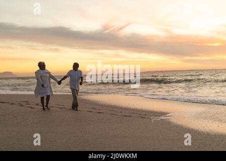 Ein Paar aus Senioren hält sich die Hände, während sie während des Sonnenuntergangs am Strand gegen den Himmel laufen Stockfoto