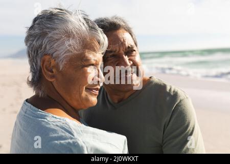 Nachdenkliches mehrrassiges Senioren-Paar, das an einem sonnigen Tag am Strand steht und zusammen wegschaut Stockfoto