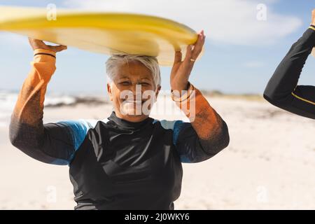 Porträt einer lächelnden, pensionierten Frau im Biracial-Alter, die am sonnigen Strand Surfbrett auf dem Kopf trägt Stockfoto