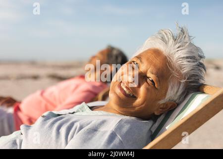 Porträt einer lächelnden älteren Birazialfrau, die sich an sonnigen Tagen mit einem Mann am Strand auf einem Stuhl entspannt Stockfoto