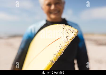 Surfbrett mit Sand, gehalten von einer lächelnden Senioren-Frau am Strand während eines sonnigen Tages Stockfoto