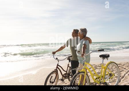 Glückliches multirassisches Seniorenpaar, das mit Fahrrädern am sonnigen Strand gegen den Himmel steht Stockfoto