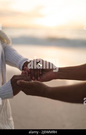 Zugeschnittenes Bild des Biracial Bräutigams, der während des Sonnenuntergangs die Hände der Braut am Strand hält Stockfoto