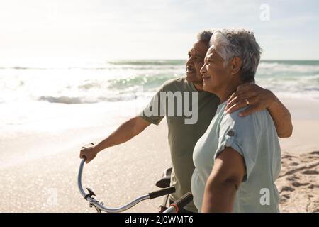 Nachdenkliches, multirassisches Seniorenpaar blickt weg, während es mit Fahrrädern am sonnigen Strand steht Stockfoto