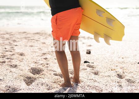 Niedriger Teil einer älteren Birazialfrau, die an sonnigen Tagen barfuß auf Sand am Strand Surfbrett trägt Stockfoto