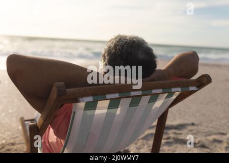 Der ältere Biracial-Mann entspannt sich während des Sonnenuntergangs mit den Händen hinter dem Kopf auf einem Klappstuhl am Strand Stockfoto